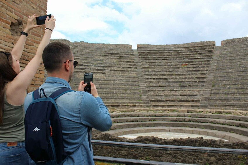 Two people taking photos of an ancient amphitheater with tiered seating. 1