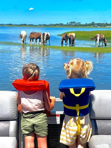Two children on a boat, wearing life jackets, looking at horses grazing by the water's edge. 3