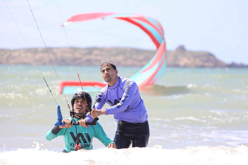 A person in a teal wetsuit holding a kiteboard, with a kite in the background. 1
