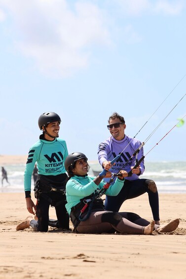 Three people on a beach with a kiteboard, one person holding the kite, and another sitting on the sand. 3