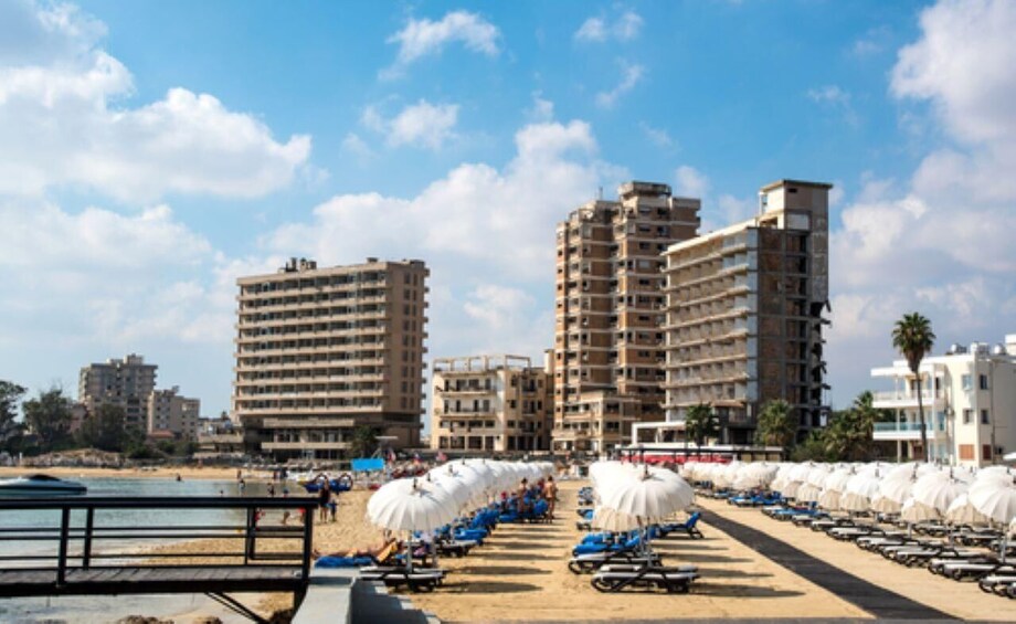 A beach with rows of sun umbrellas and lounge chairs, a wooden walkway, and several multi-story buildings in the background. 1