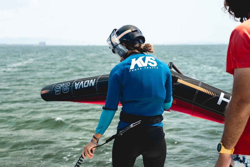A person carrying a paddleboard with 'KVS Punta Tretto' written on it, standing by the sea. 5