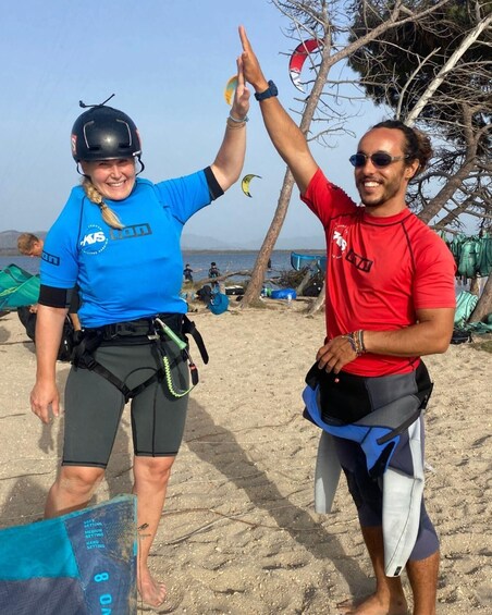 Two people on a beach, one in a blue wetsuit and helmet, the other in a red shirt, with a kite in the background. 3