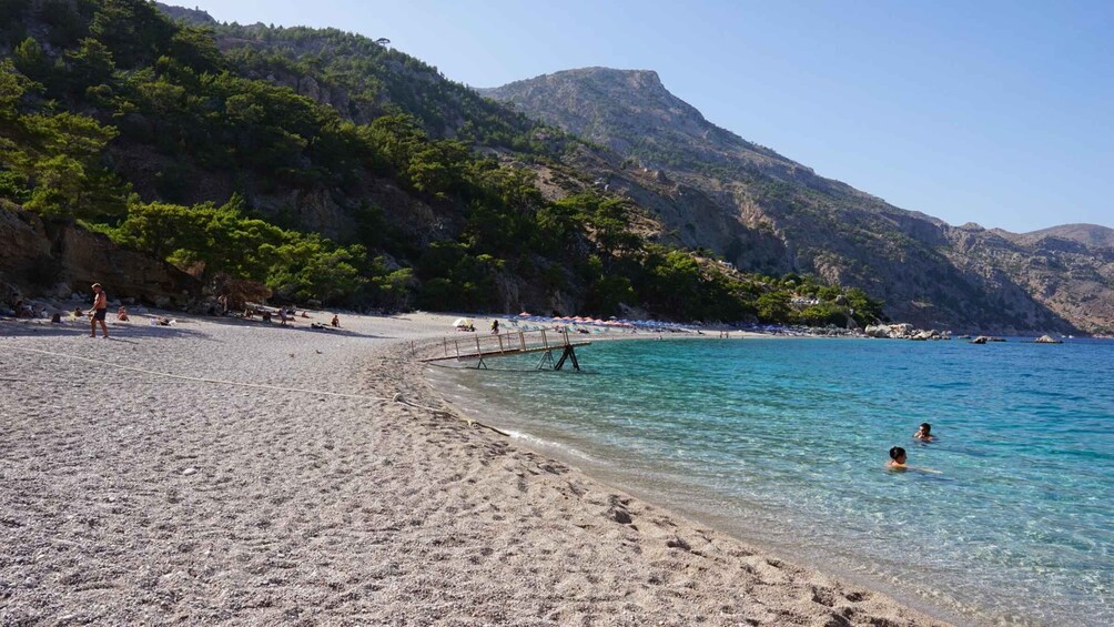 A beach with clear turquoise water, people swimming and sunbathing, and a rocky hillside in the background. 3