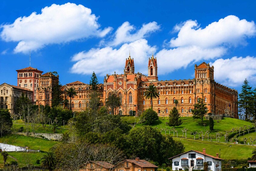 A large, historic building with multiple towers and spires, surrounded by lush greenery and a clear blue sky. 1
