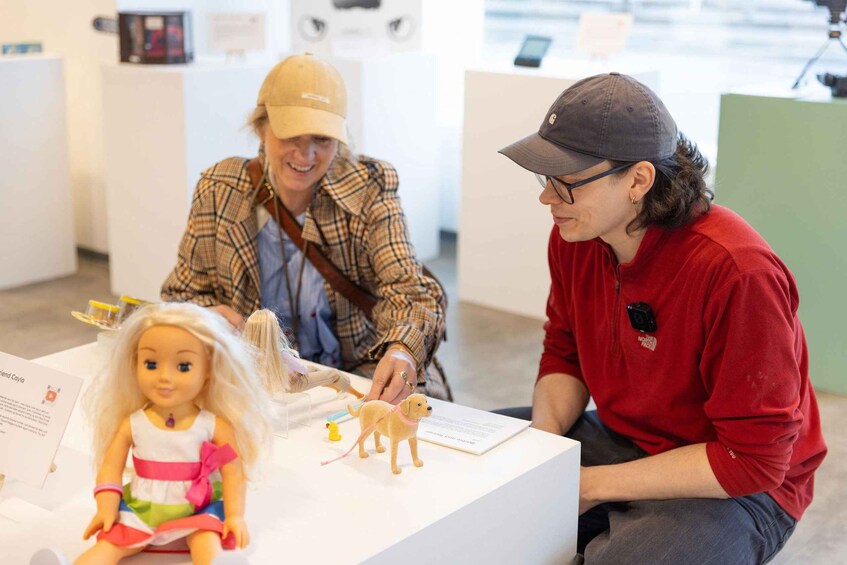 A person in a red shirt and cap sitting at a table with a doll and a small dog figurine. 1