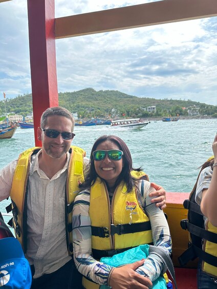 Two people on a boat wearing life jackets, with a mountainous landscape and other boats in the background. 4