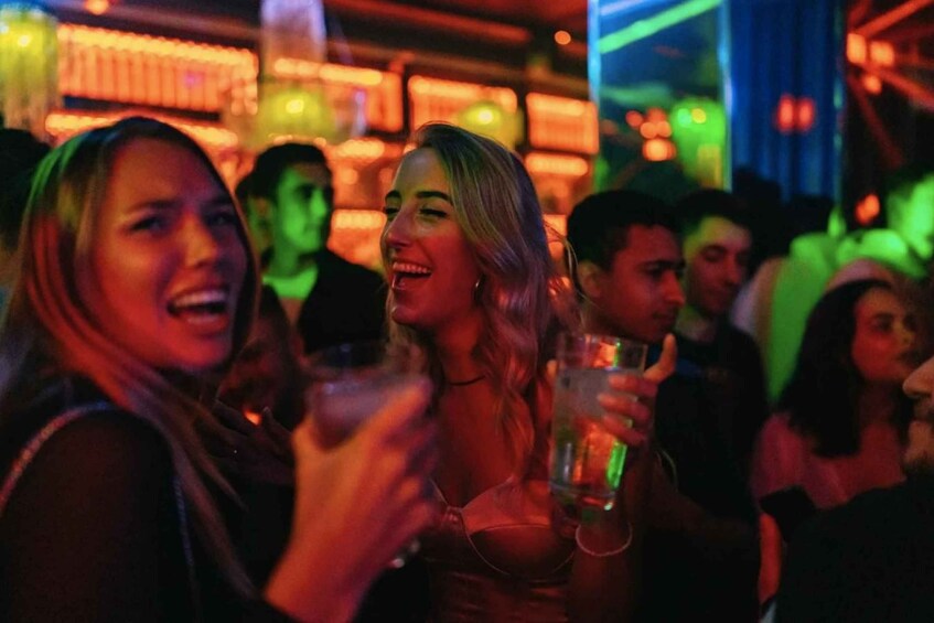 People in a dimly lit indoor setting, possibly a bar or club, with colorful lights in the background. 1