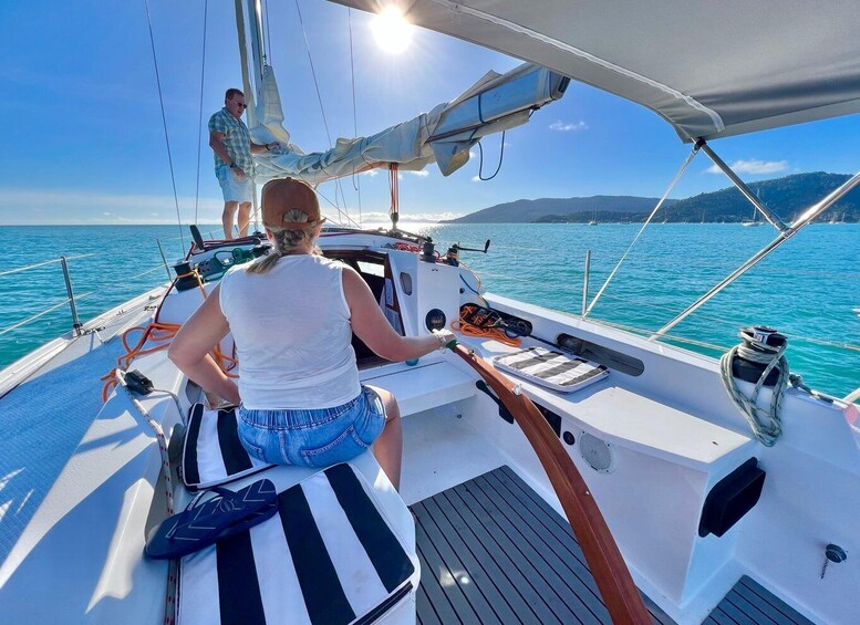 A sailboat with a woman steering, another person on the deck, and a clear blue sea with distant mountains. 5