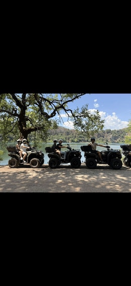 Four people on ATVs parked by a lake with a large tree and mountains in the background. 1