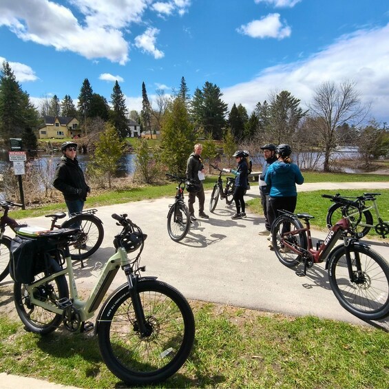 A group of cyclists on a paved path with bicycles parked nearby, surrounded by trees and a body of water. 3