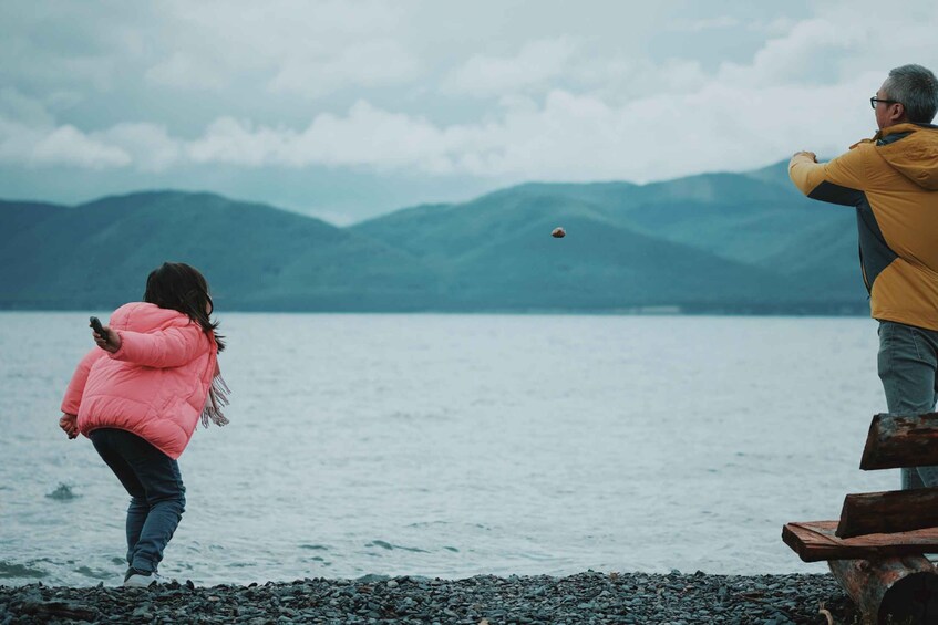 A person in a pink jacket throwing a ball, another person in a yellow jacket watching, and a lake with mountains in the background. 2