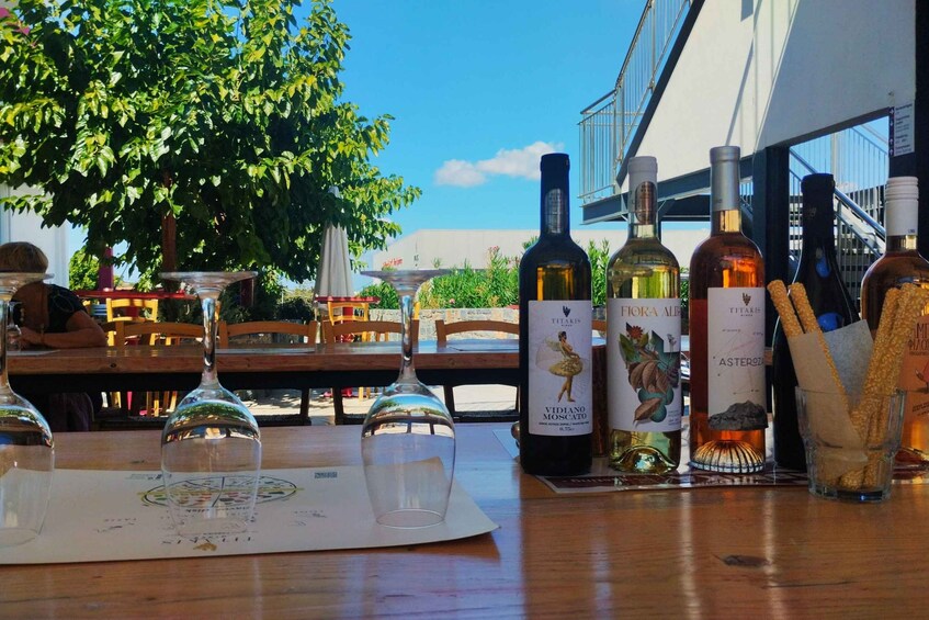 A table with wine bottles and glasses, a menu, and a view of a building and tree. 2