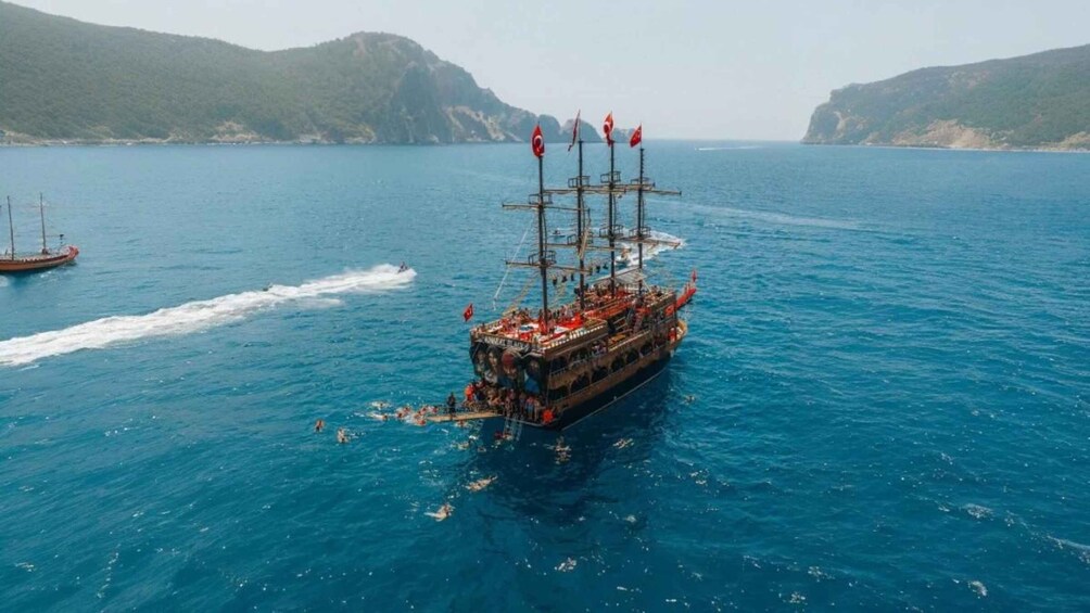 Un grand navire arborant de nombreux pavillons, naviguant sur une mer d’un bleu profond, avec une côte montagneuse en arrière-plan. 2