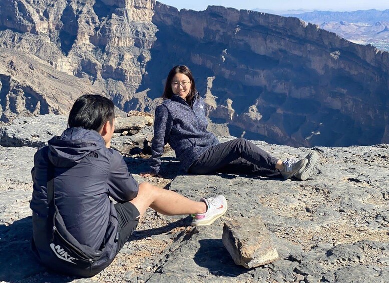 Two people sitting on a rocky mountain ledge with a canyon in the background. 1