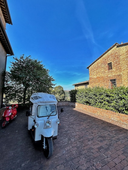 A white three-wheeled vehicle parked on a brick path, a red scooter, and a stone building with a window. 4