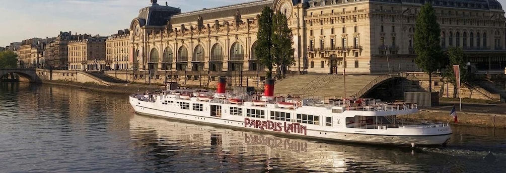 A river cruise boat named 'PARADISE' on a river with historic buildings in the background. 1
