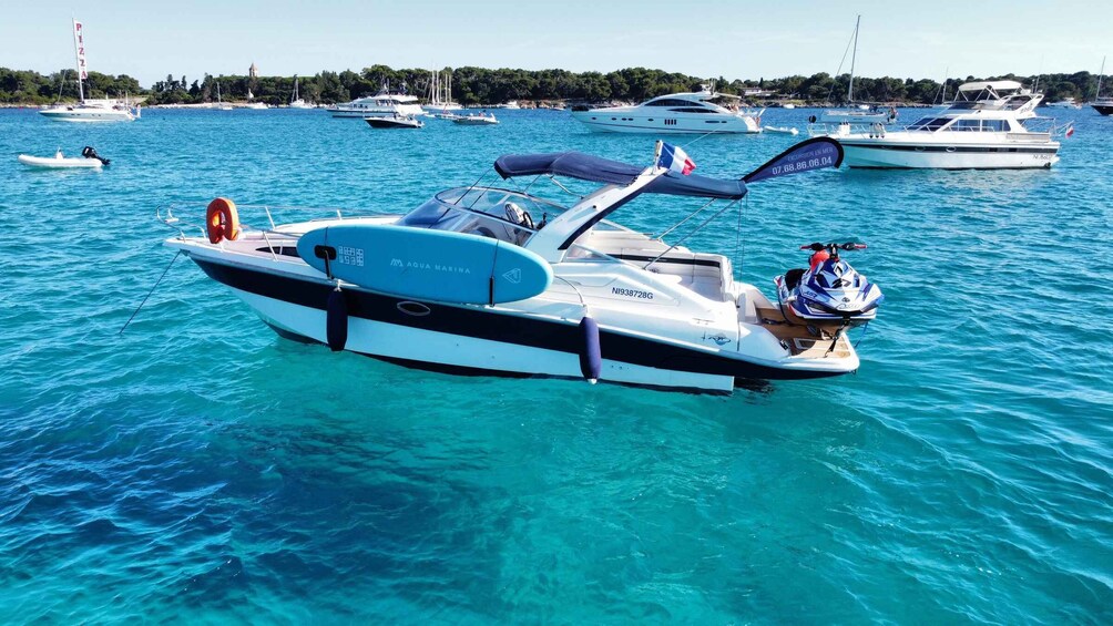 A white and blue motorboat with a canopy and a jet ski on board, anchored in a marina with other boats and a clear blue sea. 5