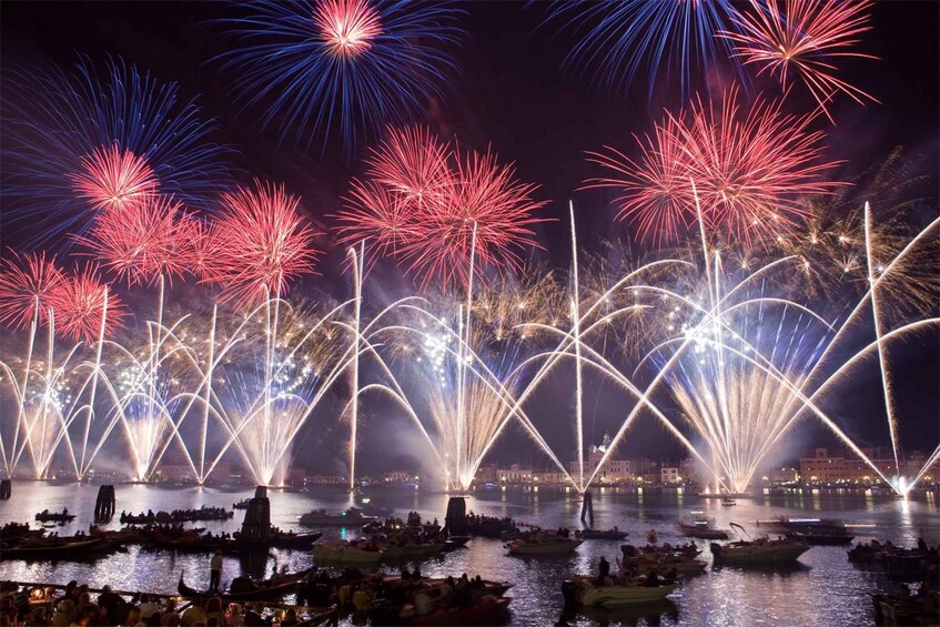 A nighttime fireworks display over a body of water with boats and a cityscape in the background. 1