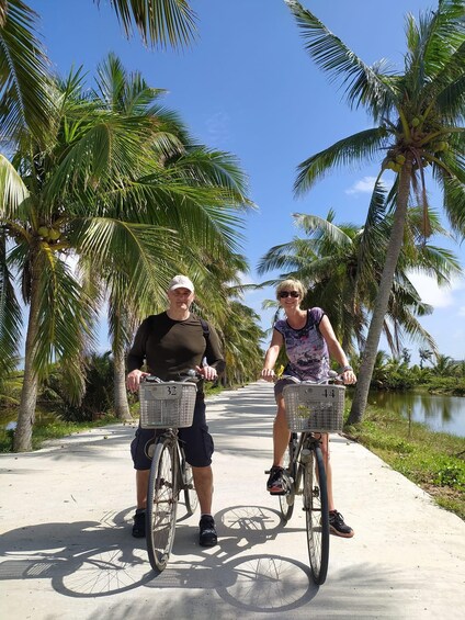 Two people on bicycles with baskets, riding on a paved path surrounded by palm trees. 5