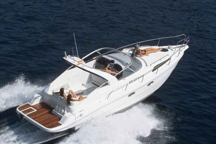 A white motorboat with a wooden deck, a person steering, and another person standing at the bow. 1