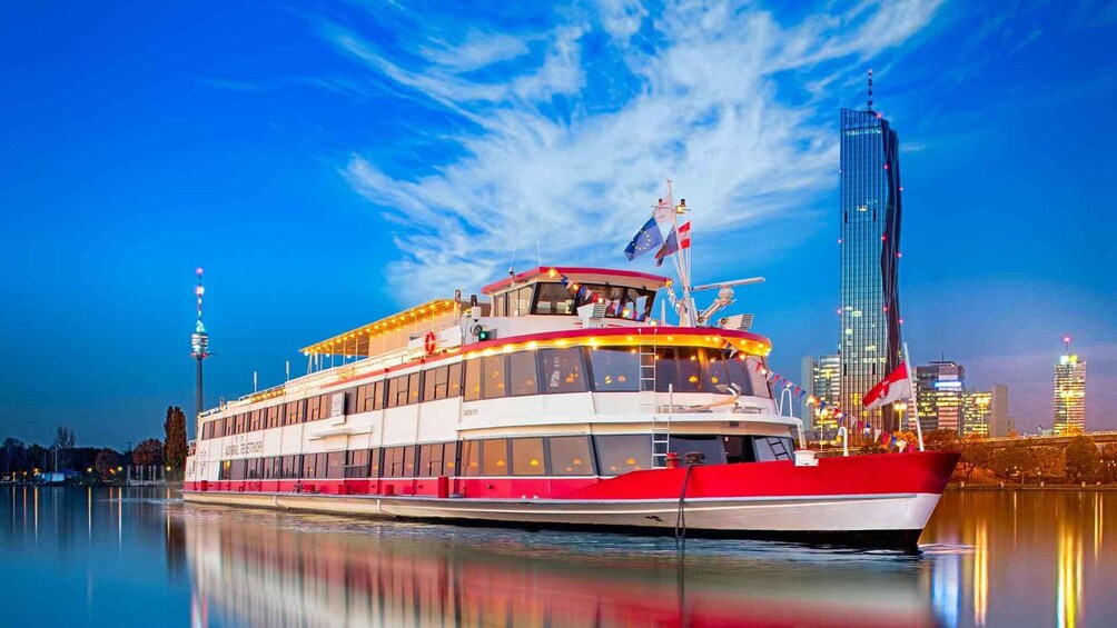 Un barco de crucero fluvial rojo y blanco con una bandera, atracado cerca de un horizonte urbano con edificios altos. 5