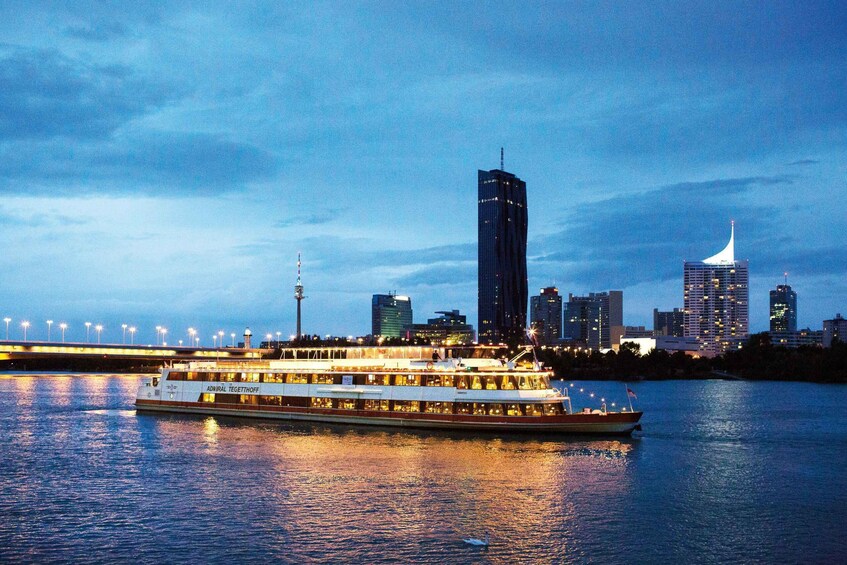 Un barco de paseo fluvial con la inscripción 'ADNARL TEGETHOFF' en su lateral, una silueta urbana con edificios modernos y un puente al fondo. 1