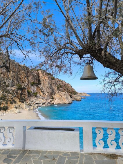 A coastal landscape with a rocky cliff, a clear blue sea, and a bell hanging from a tree branch. 2