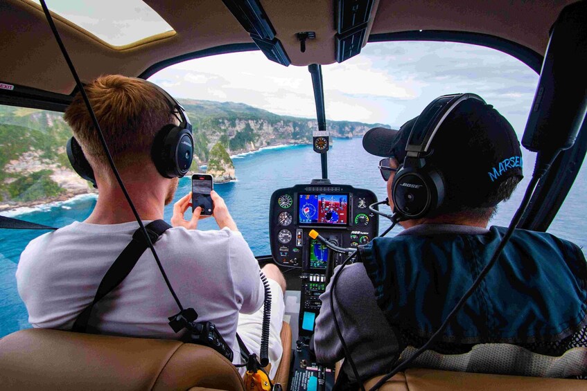 Two people in a helicopter cockpit, one using a smartphone, with a coastal landscape in the background. 1
