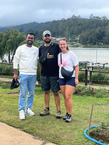 Tre persone in piedi sull'erba vicino a un lago, con alberi e case sullo sfondo. 3