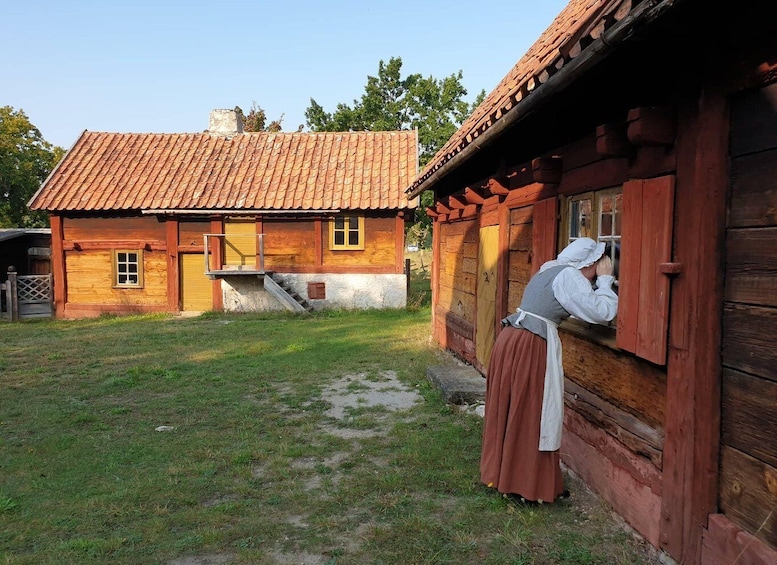 A rustic wooden house with a tiled roof, a person in traditional attire leaning out of a window, and a grassy yard. 2