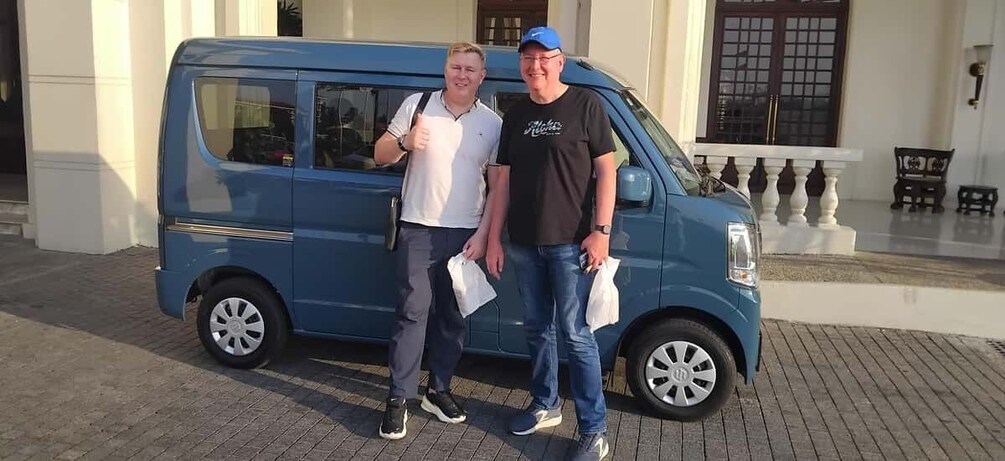 Two people standing next to a blue van in front of a building with a white facade and a balcony. 1