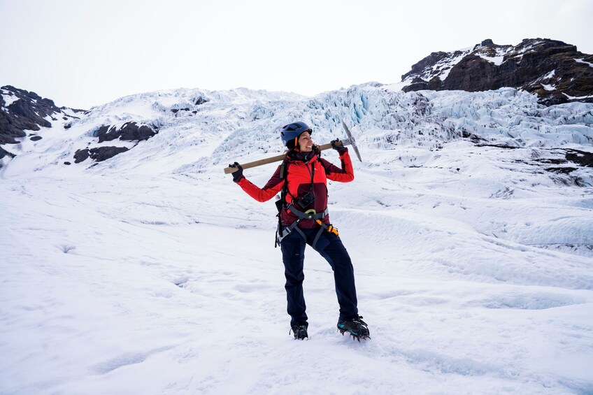 Une personne vêtue d’une veste rouge et d’un casque, tenant un bâton en bois, se tient sur un terrain enneigé avec des montagnes en arrière-plan. 5