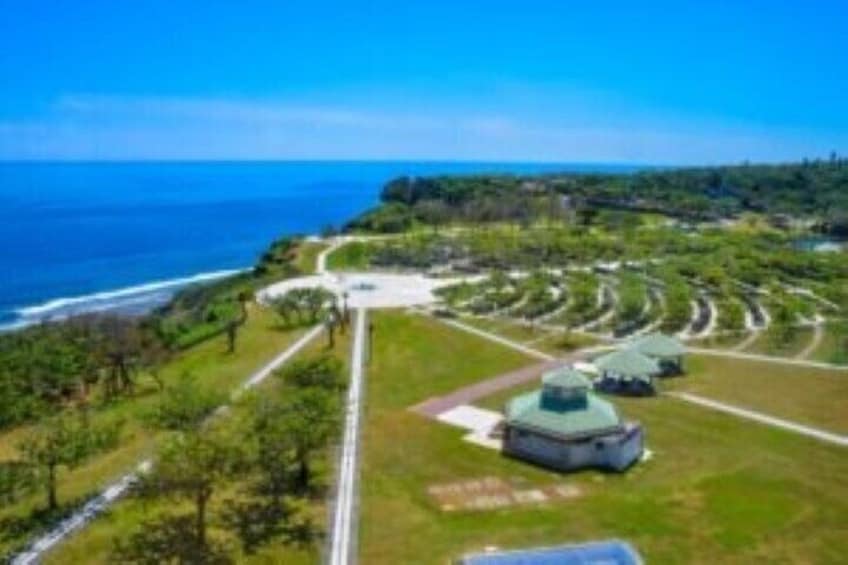 A coastal landscape with a road leading to a series of green-roofed structures, a clear blue sky, and the ocean in the background. 5