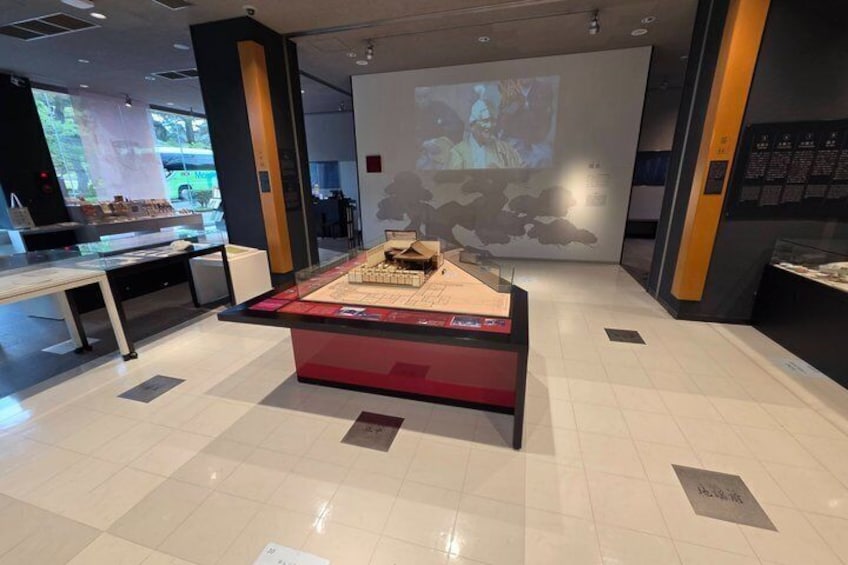 Et museumsinteriør med en central udstillingssøjle, informationsskilte og en projektorskærm i baggrunden. 1