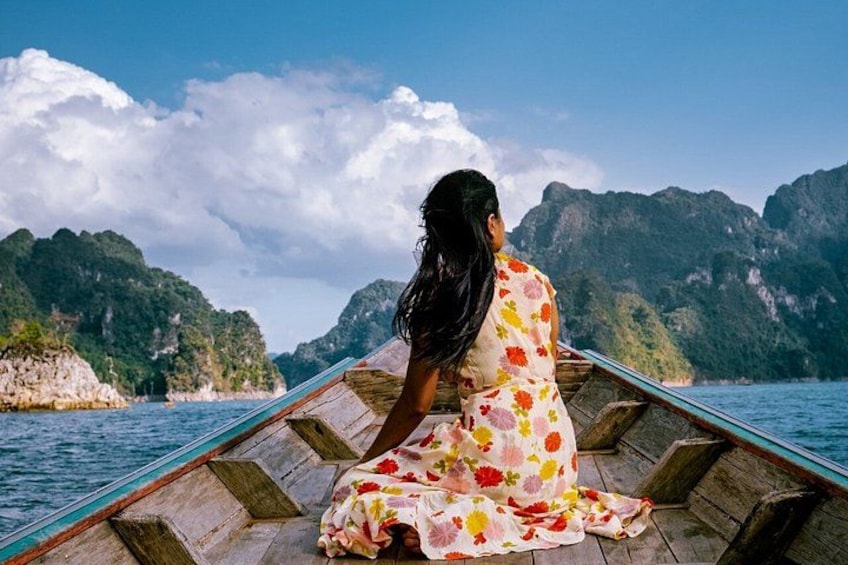 Eine Person in einem Blumenkleid sitzt auf einem hölzernen Boot vor einer bergigen Landschaft. 2