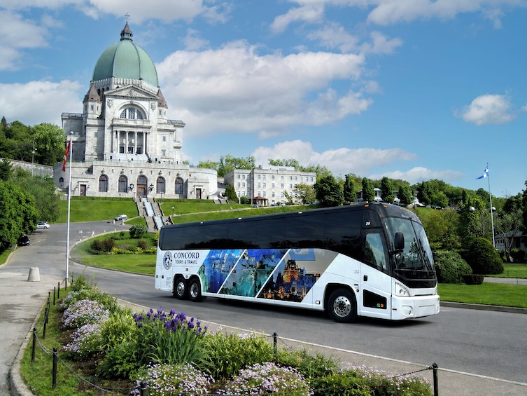 Valkoinen matkailubussi, jonka kylkeen on maalattu teksti »CONCORD TOURS & TRAVEL», on pysäköitynä suuren, kupolikattoisen rakennuksen eteen. Rakennuksen edessä on myös lipputanko, jossa liehuu lippu. 1