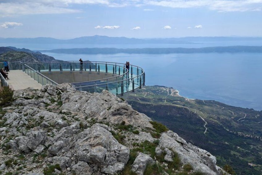 A glass observation deck overlooking a lake with rocky terrain and distant mountains. 1