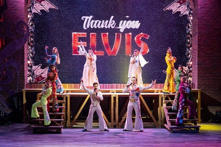 Un escenario con un telón de fondo que dice 'Thank you ELVIS', artistas con vestuario llamativo y una plataforma de madera. 5