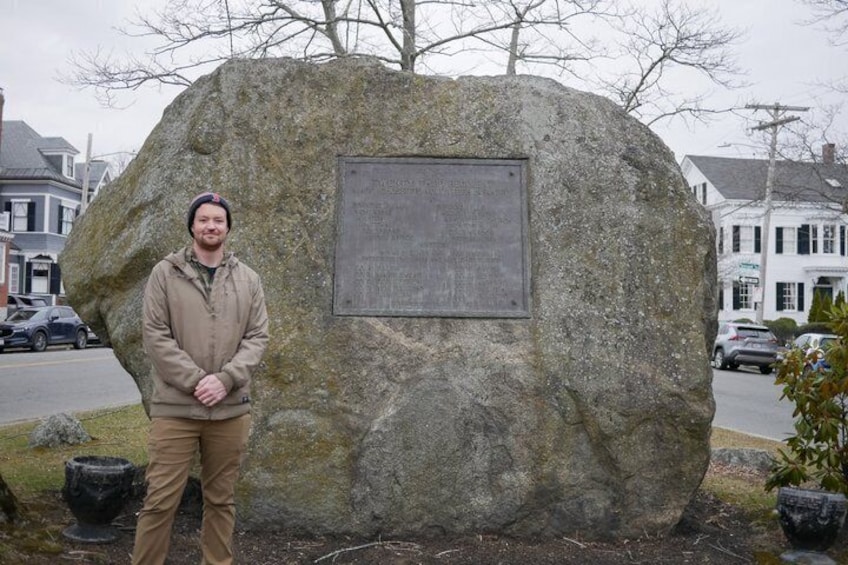 En mand står foran en stor klippe med en mindetavle, en vej og huse i baggrunden. 5