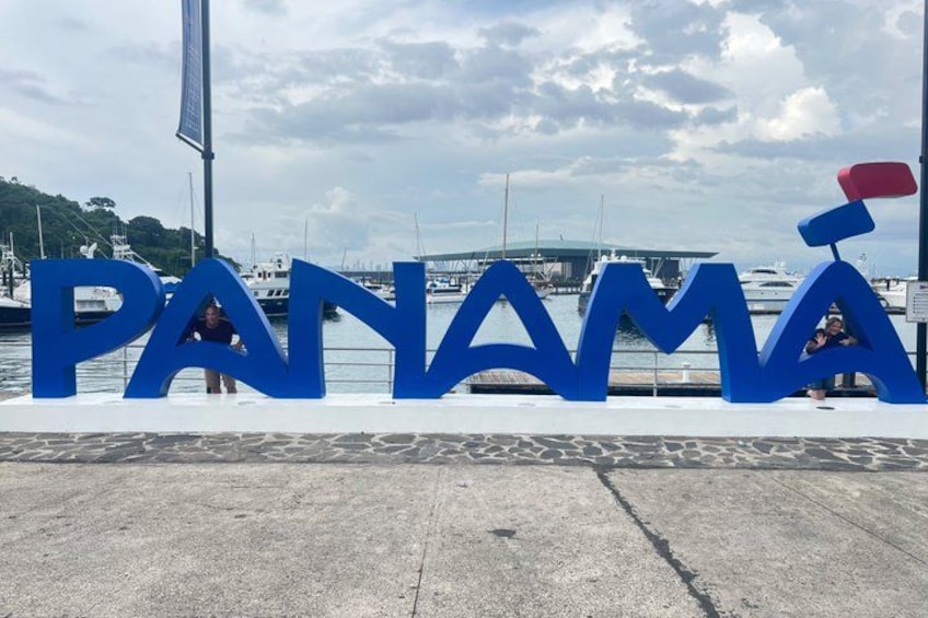 Large blue letters spelling 'PANAMÁ' with a red flag on top, set against a marina backdrop with boats and a cloudy sky. 3