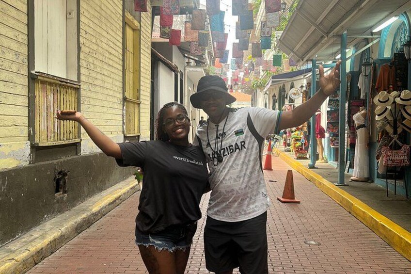 Two people posing on a brick sidewalk with shops and hanging decorations in the background. 4