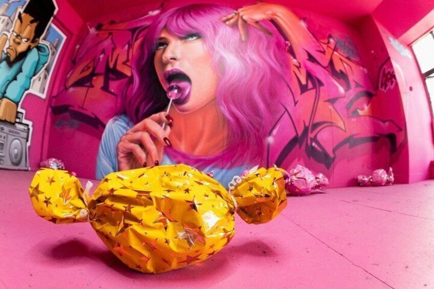En person med rosa hår, en karamellstång och ett stort, gult föremål som liknar en björn, placerat på en rosa yta med graffiti i bakgrunden. 4
