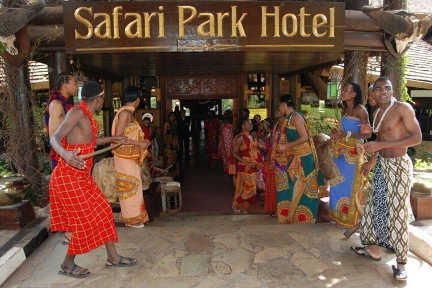 A traditional African village with people in colorful attire and a sign for Safari Park Hotel. 1