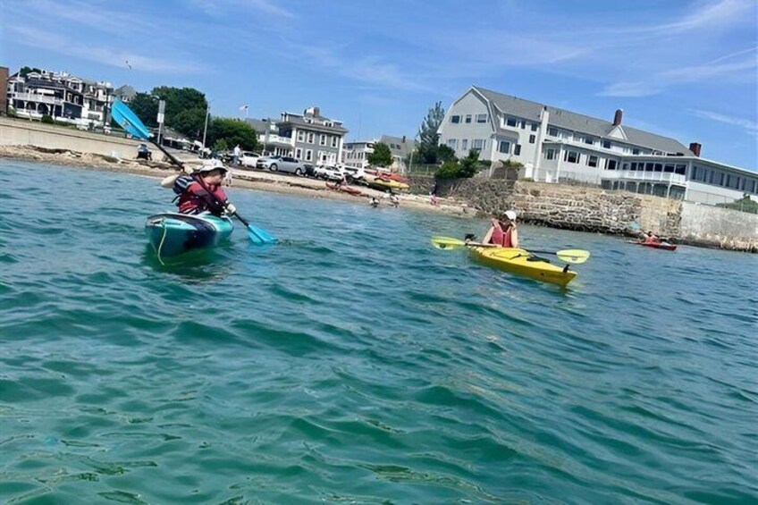 Dos kayaks sobre agua tranquila, con edificios y una playa al fondo. 5