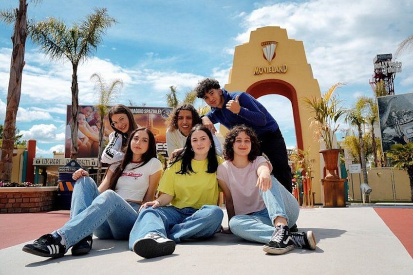 Un grupo de personas sentadas en una plataforma, con palmeras y un letrero que dice 'MOVIELAND' al fondo. 2