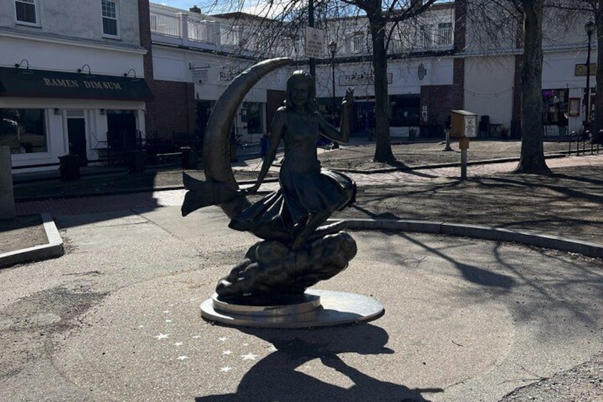 En staty som föreställer en personfigur tillsammans med en fisk, placerad på ett offentligt torg med byggnader i bakgrunden. 4