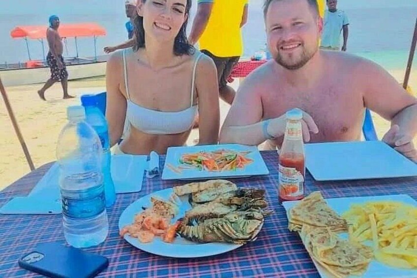 En strandscen där människor sitter vid ett bord och äter olika rätter och dricker. 2