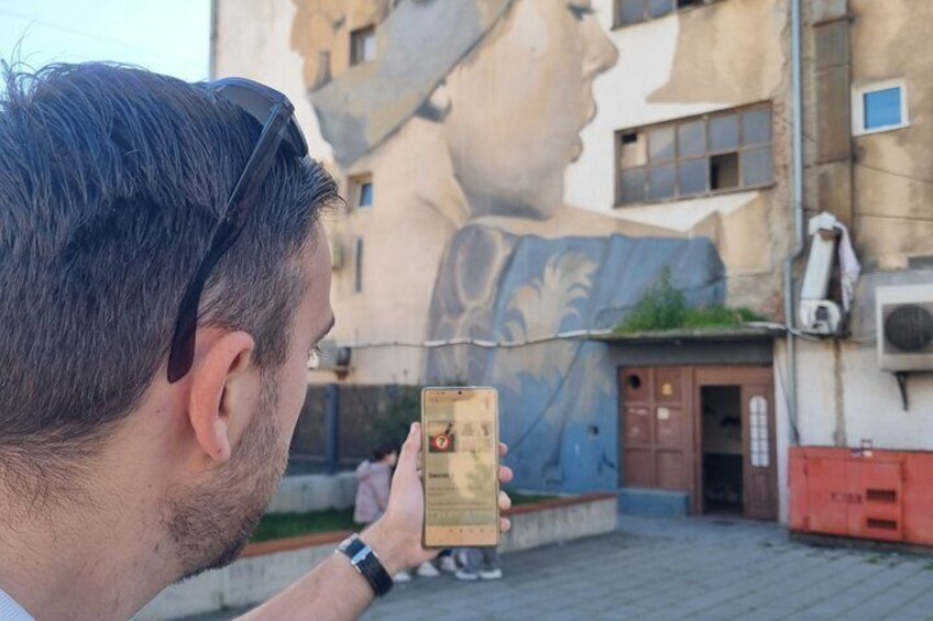 A person holding a smartphone, a mural of a woman's face on a building, and a red container on the ground. 5