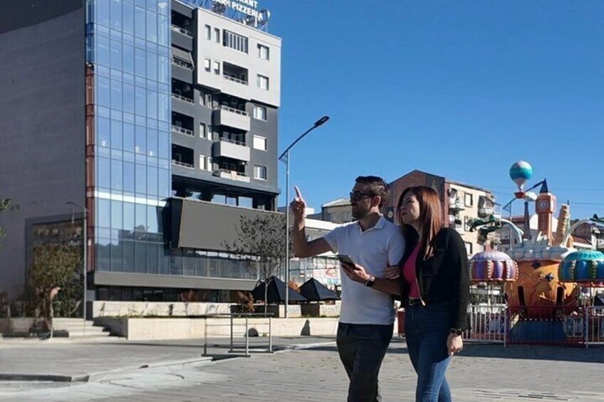 A man taking a selfie with a woman, a modern building, and a colorful amusement park in the background. 4